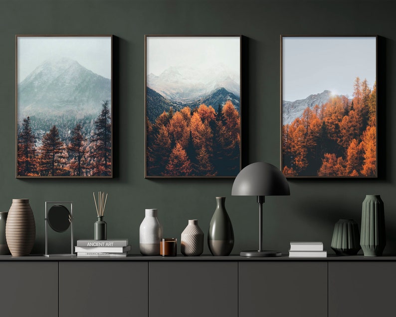 May include: Three framed landscape photographs of mountains and trees with fall foliage. The photos are hung on a dark green wall above a shelf with decorative objects.