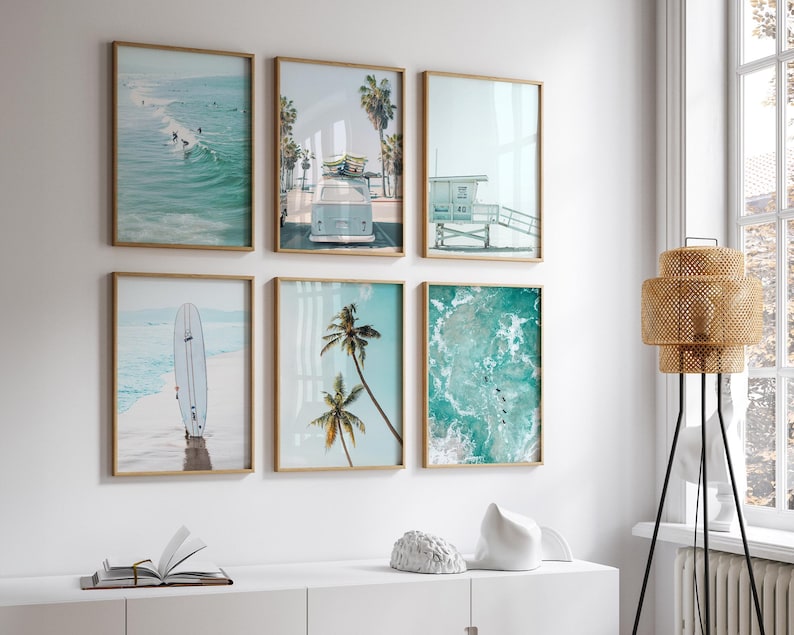 May include: Six framed prints with a beach theme. The prints feature images of a surfboard, palm trees, ocean waves, and a lifeguard stand. The prints are arranged in a grid pattern on a white wall.