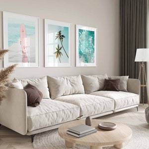 May include: A white sectional sofa with brown throw pillows in a living room with three framed beach-themed prints on the wall. The prints feature a surfboard, palm trees, and ocean waves.