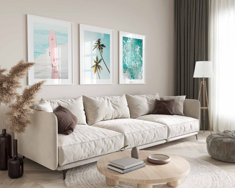May include: A white sectional sofa with brown throw pillows in a living room with three framed beach-themed prints on the wall. The prints feature a surfboard, palm trees, and ocean waves.