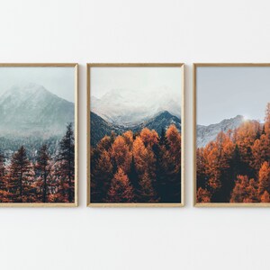 May include: Three framed prints of a mountain landscape with fall foliage. The left print shows a snow-capped mountain peak with a misty forest in the foreground. The middle print shows a forest of orange and red trees. The right print shows a mountain range with a forest of orange and red trees in the foreground.
