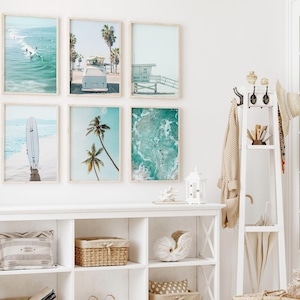 May include: A white wall with six framed prints of beach scenes. The prints feature images of surfers, palm trees, and ocean waves. The prints are arranged in a grid pattern. A white shelf with a few items on it is below the prints.