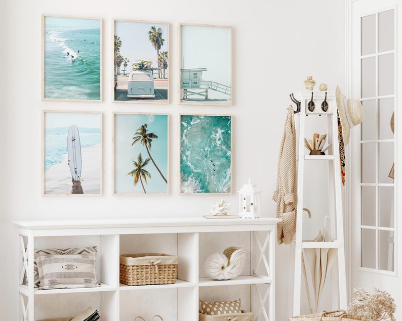 May include: A white wall with six framed prints of beach scenes. The prints feature images of surfers, palm trees, and ocean waves. The prints are arranged in a grid pattern. A white shelf with a few items on it is below the prints.