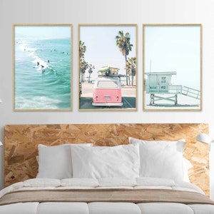 May include: Three framed prints depicting a beach scene. The first print shows a blue ocean with surfers riding waves. The second print shows a pink vintage van with surfboards on the roof driving down a street lined with palm trees. The third print shows a white lifeguard stand on a sandy beach.