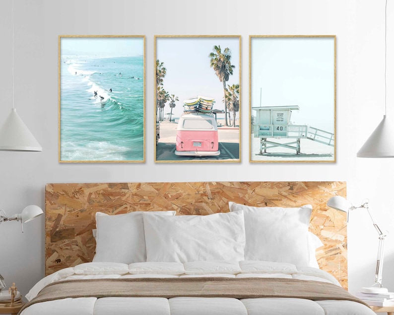 May include: Three framed prints depicting a beach scene. The first print shows a blue ocean with surfers riding waves. The second print shows a pink vintage van with surfboards on the roof driving down a street lined with palm trees. The third print shows a white lifeguard stand on a sandy beach.