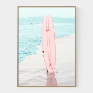 May include: A pink surfboard with the text "INT. 3" on the side, standing on a sandy beach with blue ocean water in the background.