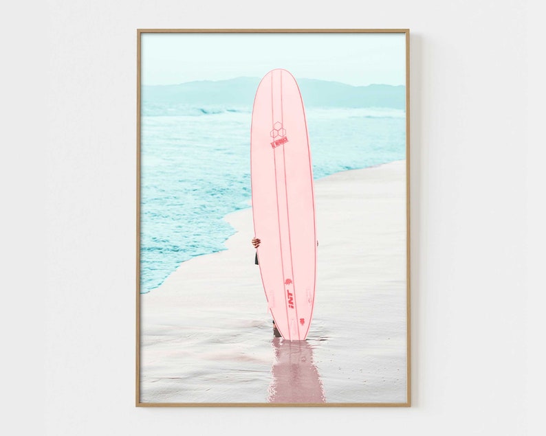 May include: A pink surfboard with the text "INT. 3" on the side, standing on a sandy beach with blue ocean water in the background.