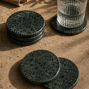 May include: A set of circular dark green stone coasters with a speckled, mottled texture. The coasters are arranged in a stack and individually, with one clear ribbed glass tumbler resting on top of one of the stone coasters.