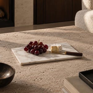 May include: A rectangular white marble serving board with grey veining, holding a cluster of dark red grapes, a small round piece of brie cheese, and a cheese knife with a dark brown handle, resting on a textured beige stone surface.