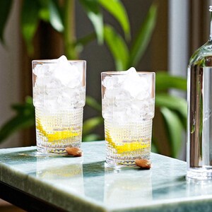 K&ouml;nnte beinhalten: Zwei klare Trinkgl&auml;ser mit einem strukturierten, facettierten Bodenmuster, jeweils gef&uuml;llt mit Eisw&uuml;rfeln und einer Scheibe gelber Zitrone. Neben jedem Glas liegt eine braune Mandel auf der Oberfl&auml;che, rechts steht eine klare Glasflasche.