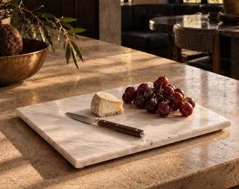 May include: A rectangular white marble serving board featuring a wedge of soft cheese, a cluster of dark red grapes, and a small knife with a dark brown handle. The board has a smooth, polished surface with subtle grey veining throughout the stone.