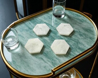 May include: Four white hexagonal marble coasters arranged on an oval green marble bar cart with a gold metal frame. Two clear glass tumblers are placed on the top surface, while a small bowl of fruit and a glass sit on the lower shelf.