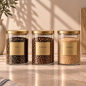 May include: Three clear glass cylindrical canisters with brushed gold lids. Each jar features a square gold label with black text reading TEA, COFFEE, or SUGAR, and the words 100% NATURAL. The jars are filled with loose tea leaves, coffee beans, and sugar.