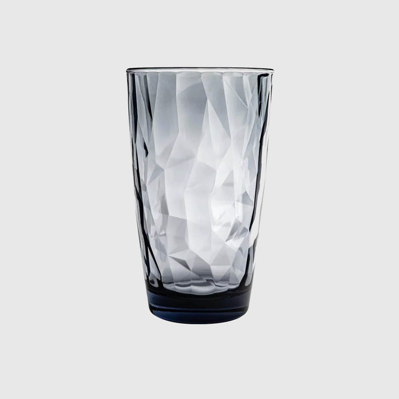 May include: A tall, cylindrical drinking glass made of translucent, smoky gray glass. The exterior surface features a faceted, geometric texture with numerous angular planes that catch the light, creating a complex, multi-faceted appearance.