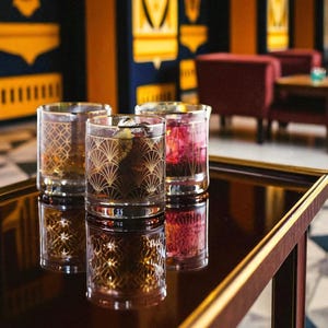 Puede incluir: Tres vasos de cristal transparente con un patr&oacute;n geom&eacute;trico de abanico en color dorado. Cada vaso contiene un l&iacute;quido de diferente color, incluyendo &aacute;mbar, marr&oacute;n oscuro y rojo intenso, con cubitos de hielo visibles dentro de los recipientes.