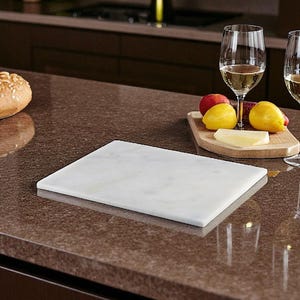 May include: A rectangular white marble cutting board sits on a brown countertop. The board is smooth and reflective. A wooden serving board with fruit and cheese, and two wine glasses are in the background. A loaf of bread is on the left.