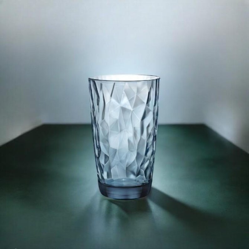 May include: A clear, colorless drinking glass featuring a faceted, geometric texture across its exterior surface. The glass has a cylindrical shape that tapers slightly toward the base, creating a multi-dimensional appearance with light reflections.
