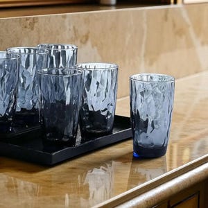 May include: A set of five blue drinking glasses with a faceted, geometric texture. Four of the glasses are arranged on a rectangular black tray, while one glass stands separately to the right. The glass surfaces feature a crinkled, angular pattern.
