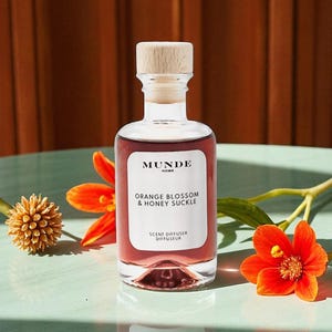 May include: A clear glass scent diffuser bottle with a light wooden stopper, filled with a reddish-pink liquid. The white label reads MUNDE HOME, ORANGE BLOSSOM & HONEY SUCKLE, SCENT DIFFUSER DIFFUSEUR in black text. Bright orange flowers sit nearby.