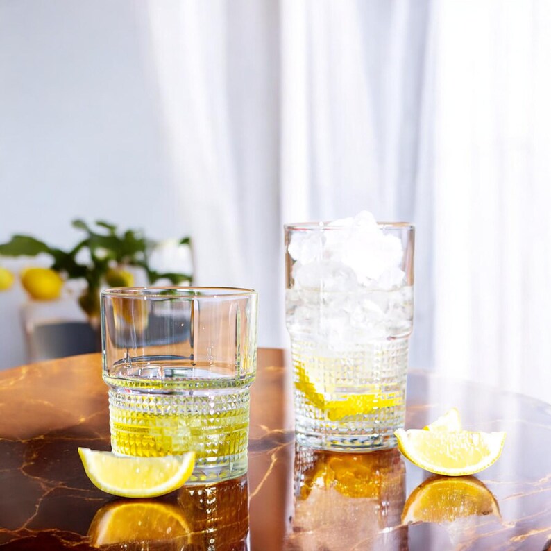 K&ouml;nnte beinhalten: Zwei klare Trinkgl&auml;ser mit einem strukturierten Rautenmuster in der unteren H&auml;lfte. Ein Glas ist mit Eisw&uuml;rfeln und einer Zitronenscheibe gef&uuml;llt, w&auml;hrend das andere eine gelbe Fl&uuml;ssigkeit und eine Zitronenscheibe enth&auml;lt. Zwei Zitronenspalten liegen daneben.
