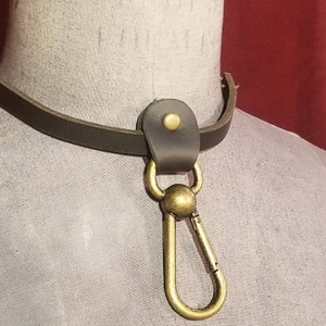 Peut inclure: Un collier en cuir marron avec un fermoir en laiton et un anneau métallique décoratif. Le collier est sur un mannequin.