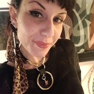 Op de afbeelding: Een zwarte leren choker ketting met een grote goudkleurige ringhanger. Een kleine bedel hangt aan de ring. Een sjaal met luipaardprint is om de nek gedrapeerd.