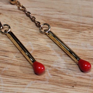 May include: A pair of gold-colored earrings with a matchstick design. Each earring features a long, rectangular gold-colored body with a small, rounded red tip. The earrings are connected by a delicate gold chain.