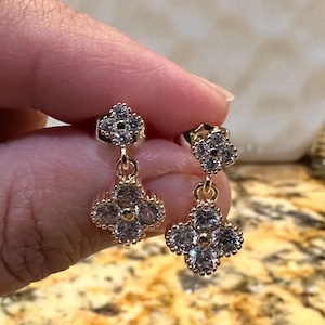 May include: A pair of gold-toned drop earrings featuring a four-leaf clover motif. Each earring consists of two clover-shaped charms connected by a small jump ring, with each clover encrusted with clear, sparkling stones and a beaded metal edge.