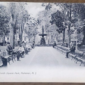 May include: A black and white photograph of a fountain in a park. The fountain is surrounded by benches and trees. The text "Fountain, Church Square Park, Hoboken, N. J." is printed at the bottom of the image.