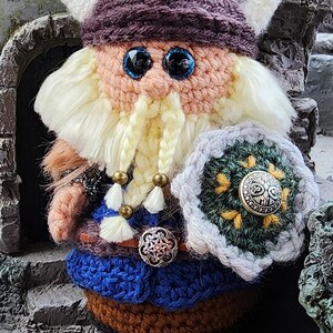 May include: A handcrafted crochet Viking doll with a purple hat, white horns, and a long, braided beard. The doll wears a blue and brown outfit and holds a shield with a decorative button. The doll has large blue eyes.