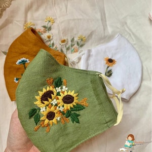 May include: Three handmade face masks with embroidered sunflower designs. The masks are made of different fabrics, including green, white, and mustard yellow.