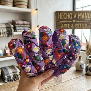 Puede incluir: Una mano sostiene varias diademas trenzadas. Las diademas son de color lavanda con patrones florales bordados en varios colores. El fondo incluye un letrero de madera que dice "HECHO A MANO ARTE & ESTILO."