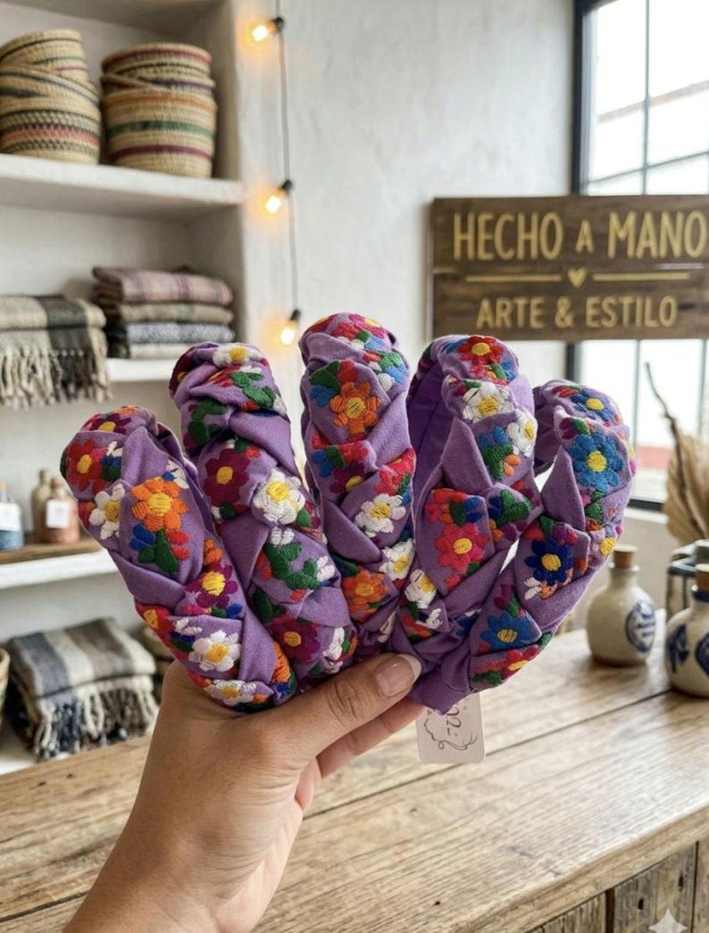Puede incluir: Una mano sostiene varias diademas trenzadas. Las diademas son de color lavanda con patrones florales bordados en varios colores. El fondo incluye un letrero de madera que dice "HECHO A MANO ARTE & ESTILO."