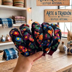 Puede incluir: Una mano sostiene varias diademas negras con bordados florales coloridos. Las diademas tienen un dise&ntilde;o trenzado y est&aacute;n adornadas con flores rojas, naranjas, amarillas, azules y verdes. El fondo incluye estantes con cestas tejidas y el letrero "ARTE & TRADICI&Oacute;N".