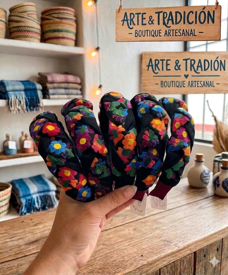 Puede incluir: Una mano sostiene varias diademas negras con bordados florales coloridos. Las diademas tienen un dise&ntilde;o trenzado y est&aacute;n adornadas con flores rojas, naranjas, amarillas, azules y verdes. El fondo incluye estantes con cestas tejidas y el letrero "ARTE & TRADICI&Oacute;N".