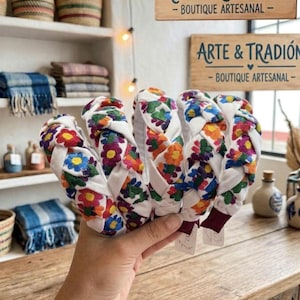 Puede incluir: Varias diademas blancas con un dise&ntilde;o trenzado, adornadas con flores bordadas coloridas en rojo, naranja, azul y verde. El fondo incluye estantes con cestas tejidas, textiles y letreros que dicen "ARTE & TRADICI&Oacute;N - BOUTIQUE ARTESANAL".