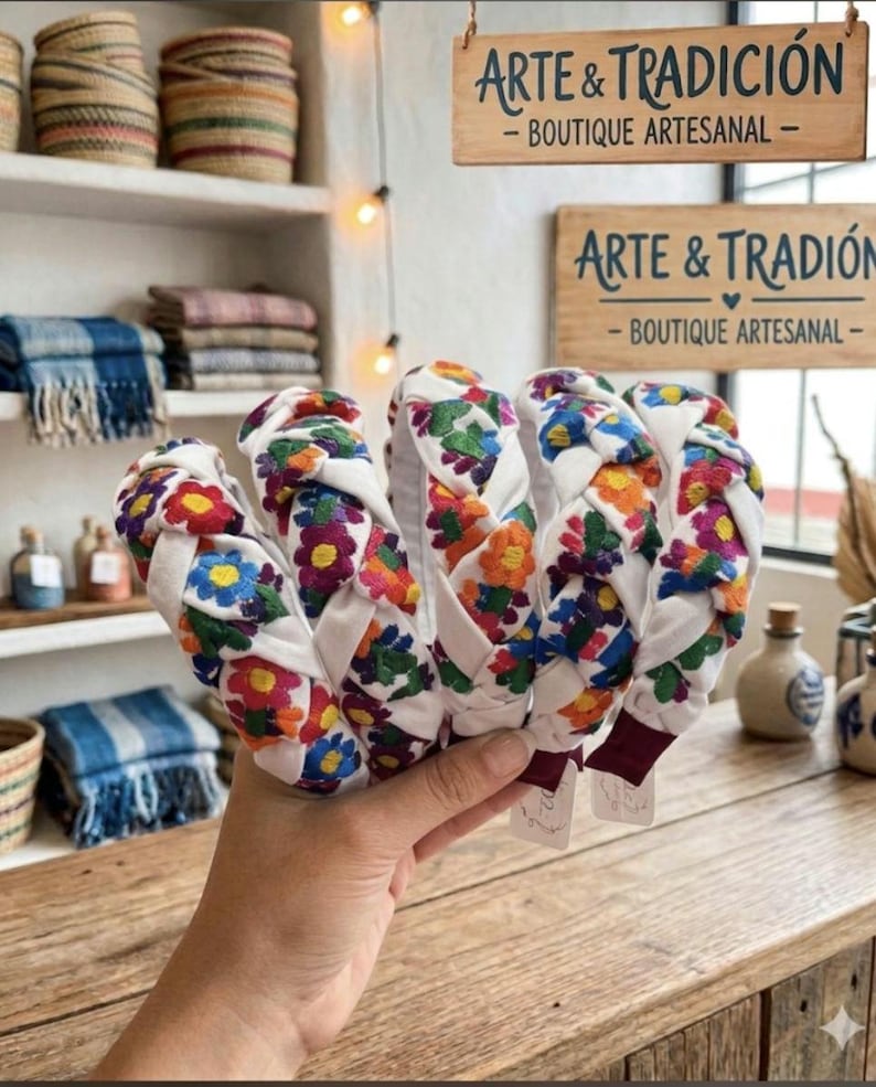 Puede incluir: Varias diademas blancas con un dise&ntilde;o trenzado, adornadas con flores bordadas coloridas en rojo, naranja, azul y verde. El fondo incluye estantes con cestas tejidas, textiles y letreros que dicen "ARTE & TRADICI&Oacute;N - BOUTIQUE ARTESANAL".