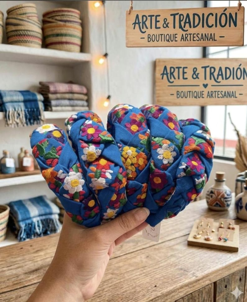 Puede incluir: Diademas de tela azul con bordados florales coloridos. Las diademas se exhiben sobre una superficie de madera, con un letrero que dice "ARTE & TRADICI&Oacute;N - BOUTIQUE ARTESANAL" al fondo. Se ven otros art&iacute;culos artesanales.