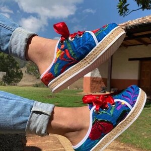 以下が含まれることがあります： カラフルな花の刺繍と赤いリボンレースが付いた青いエスパドリーユシューズ。靴は織りのジュートソールと白いトリムが付いています。青いジーンズを履いています。