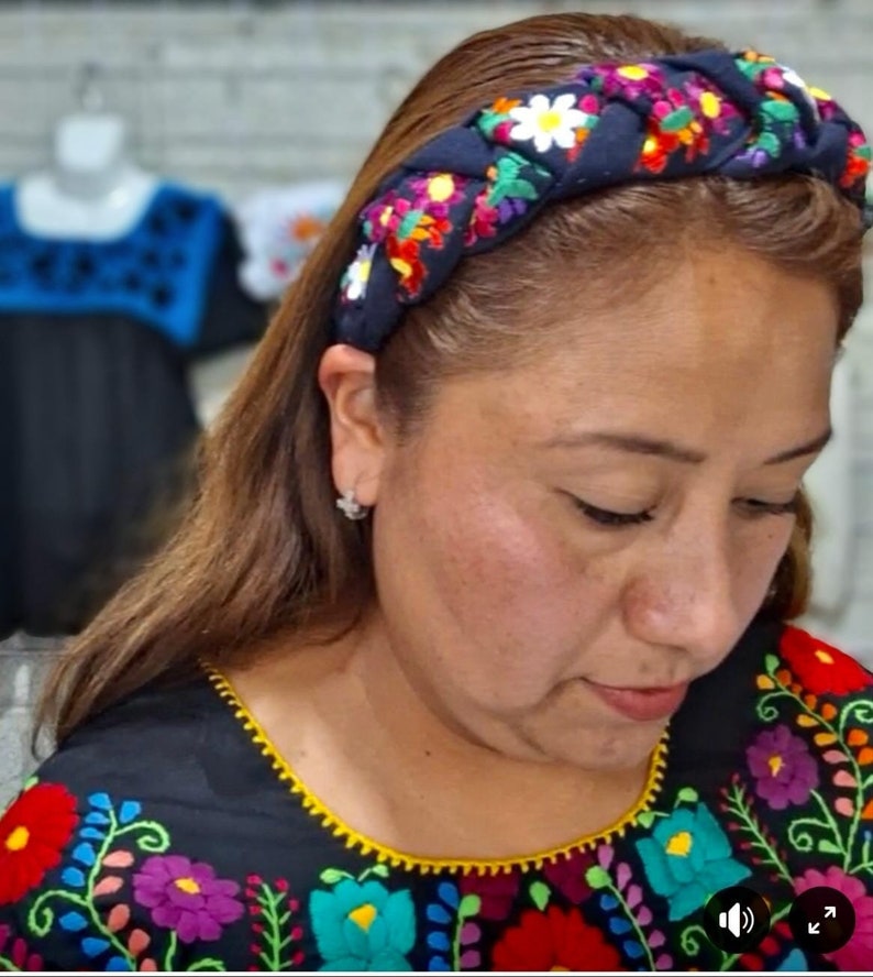Puede incluir: Una diadema trenzada con base azul oscuro, adornada con flores bordadas coloridas. La diadema presenta margaritas blancas y otros patrones florales vibrantes. La mujer de la imagen lleva un top negro bordado.
