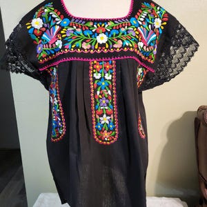 以下が含まれることがあります： カラフルな花柄刺繍が施された黒いチュニック。スクエアネックと袖はピンクのステッチと黒いレースでトリミングされています。チュニックは前面に垂直な刺繍の花柄が特徴です。