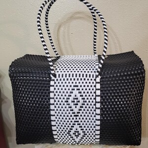 May include: A black and white woven tote bag with a rectangular shape. The bag features a central white panel with a geometric pattern, flanked by black woven sides. The handles are black and white striped.