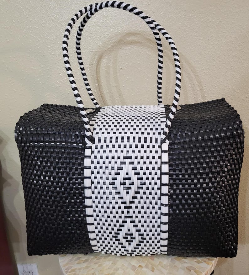 May include: A black and white woven tote bag with a rectangular shape. The bag features a central white panel with a geometric pattern, flanked by black woven sides. The handles are black and white striped.