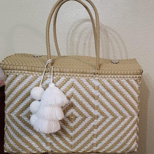 May include: Woven tote bag with a geometric diamond pattern in beige and white. The bag has two woven handles and is adorned with white pom-poms and tassels. The bag is rectangular and appears to be made of a durable material.