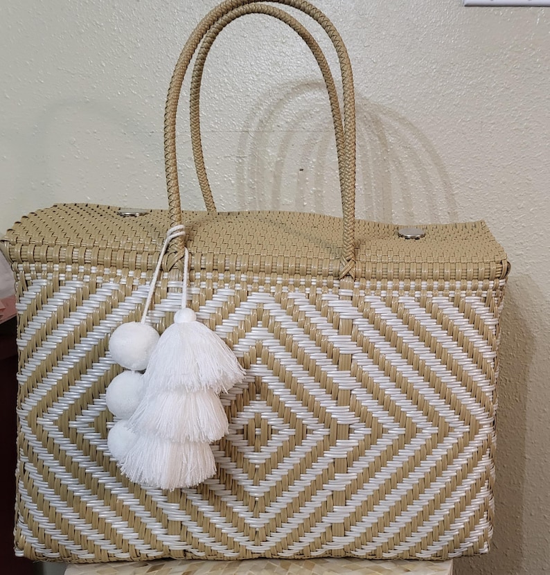 May include: Woven tote bag with a geometric diamond pattern in beige and white. The bag has two woven handles and is adorned with white pom-poms and tassels. The bag is rectangular and appears to be made of a durable material.