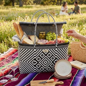 May include: A black and white woven picnic basket filled with food and drinks, including bread, cheese, grapes, and wine. The basket sits on a colorful blanket with wine glasses and plates. A wooden cutting board and knife are also present.