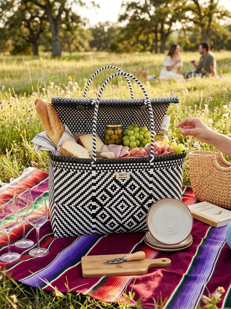 May include: A black and white woven picnic basket filled with food and drinks, including bread, cheese, grapes, and wine. The basket sits on a colorful blanket with wine glasses and plates. A wooden cutting board and knife are also present.