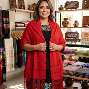 May include: A vibrant red shawl with intricate woven detailing and long fringe along the bottom edge, worn over a black dress featuring colorful floral embroidery around the neckline and hem, displayed in a shop with shelves of textiles.