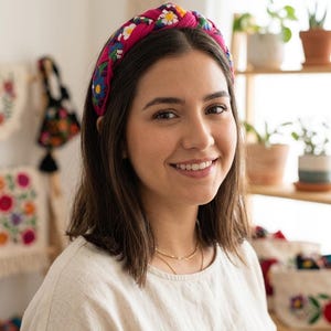 Puede incluir: Una diadema rosa brillante adornada con un colorido estampado floral. La diadema, anudada en la parte superior, es usada por una persona. El texto en la parte inferior dice: COLECCI&Oacute;N ARTESANAL - BORDADO A MANO.