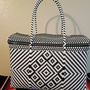 May include: Black and white woven tote bag with a geometric diamond pattern. The bag has two arched handles and a rectangular shape. The bag is sitting on a colorful striped textile.